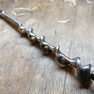 Puede incluir: Una broca de metal plateado con un diseño en espiral. La broca está sobre una superficie de madera.