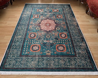 May include: A teal and orange patterned rug with a white fringe. The rug is laid out on a hardwood floor in a living room with two red velvet armchairs and a small table with a vase of flowers.