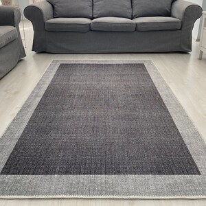 Puede incluir: Una alfombra gris con un borde blanco. La alfombra está en una sala de estar con un sofá gris y paredes blancas.