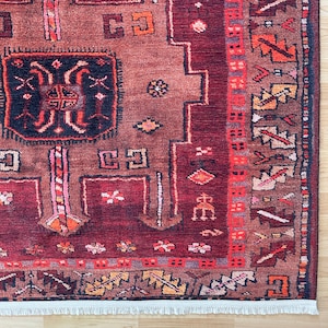 Puede incluir: Primer plano de una alfombra estampada de colores intensos. La alfombra presenta un fondo burdeos oscuro con intrincados diseños geométricos en tonos marrón, naranja y crema. El borde tiene un patrón detallado con una franja crema.