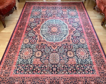 May include: A traditional Persian rug with a detailed floral and geometric pattern in shades of pink, blue, and black. The rug has a fringe border and is laid out on a wooden floor.