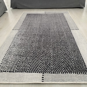 Pu&ograve; includere: Un tappeto a chevron bianco e nero con un bordo bianco. Il tappeto &egrave; steso su un pavimento bianco davanti a un divano grigio.
