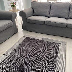 Pu&ograve; includere: Un tappeto a chevron bianco e nero con un bordo bianco. Il tappeto &egrave; in un soggiorno con un divano grigio e una gabbia per uccelli bianca.
