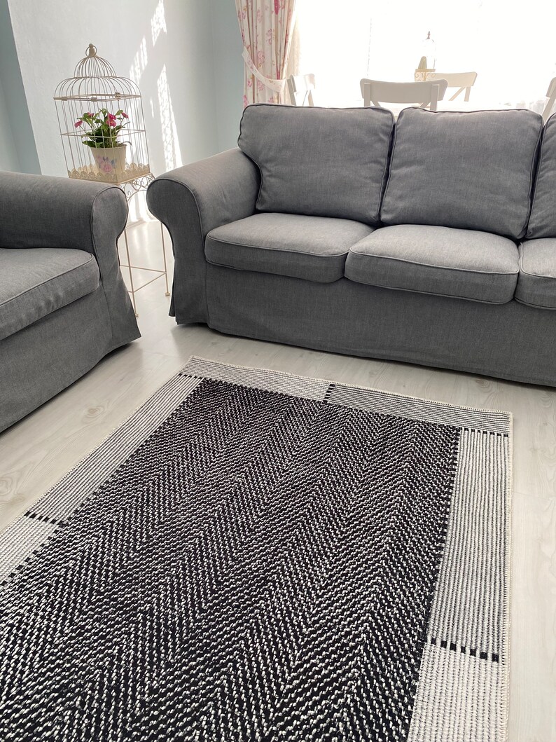 Pu&ograve; includere: Un tappeto a chevron bianco e nero con un bordo bianco. Il tappeto &egrave; in un soggiorno con un divano grigio e una gabbia per uccelli bianca.