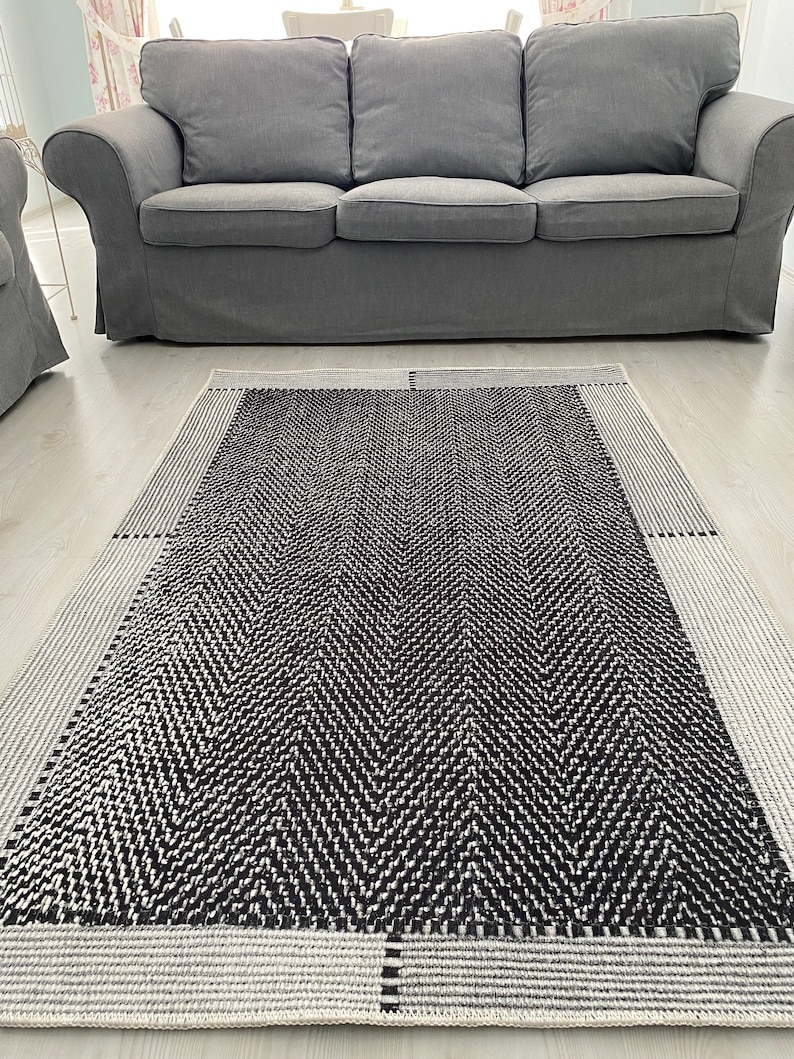 Pu&ograve; includere: Un tappeto a chevron bianco e nero con un bordo bianco. Il tappeto &egrave; steso su un pavimento bianco davanti a un divano grigio.