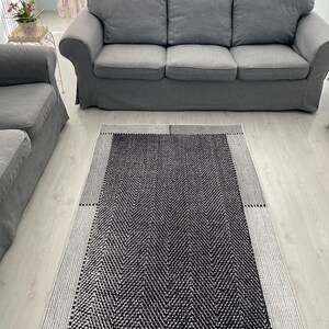 Pu&ograve; includere: Un tappeto geometrico bianco e nero con un motivo a spina di pesce. Il tappeto &egrave; su un pavimento in legno chiaro davanti a un divano grigio.