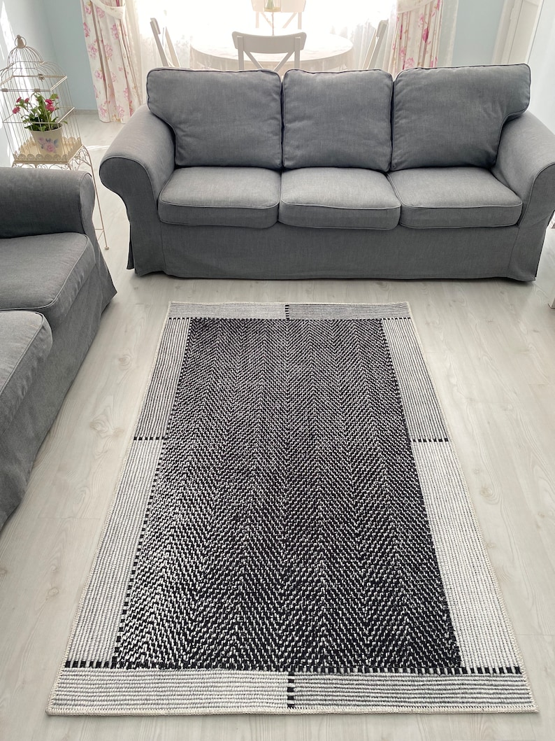 Pu&ograve; includere: Un tappeto geometrico bianco e nero con un motivo a spina di pesce. Il tappeto &egrave; su un pavimento in legno chiaro davanti a un divano grigio.