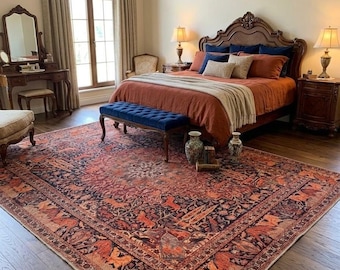 May include: A large rectangular area rug featuring a complex pattern of orange, rust, and dark blue floral and animal motifs. The rug covers a significant portion of the dark wood floor in a bedroom, positioned beneath a bed with rust-colored bedding.