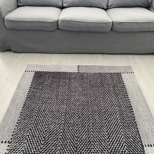 Pu&ograve; includere: Un tappeto a chevron bianco e nero con un bordo grigio. Il tappeto &egrave; steso su un pavimento grigio chiaro davanti a un divano grigio.