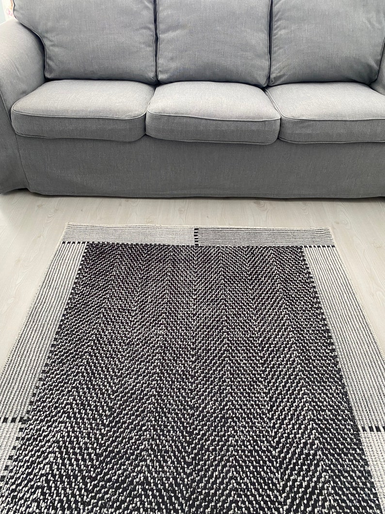 Pu&ograve; includere: Un tappeto a chevron bianco e nero con un bordo grigio. Il tappeto &egrave; steso su un pavimento grigio chiaro davanti a un divano grigio.