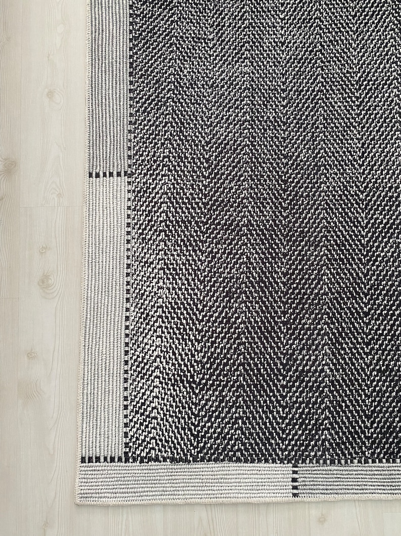 Pu&ograve; includere: Tappeto a chevron bianco e nero con bordo bianco. Il tappeto ha un design geometrico con una texture sottile.