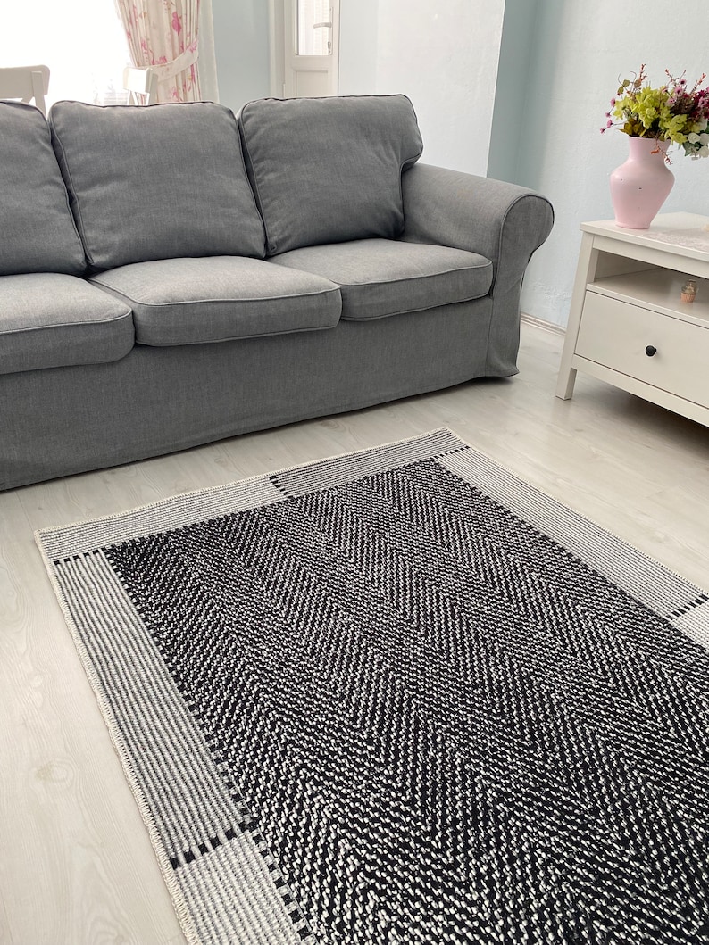 Pu&ograve; includere: Un tappeto con motivo bianco e nero con un bordo bianco. Il tappeto &egrave; steso su un pavimento in legno chiaro davanti a un divano grigio.