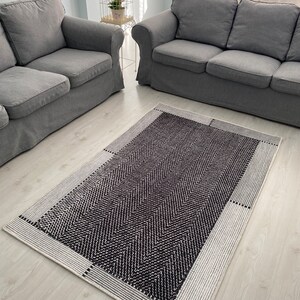 Pu&ograve; includere: Un tappeto a chevron nero e bianco con un bordo bianco. Il tappeto &egrave; posizionato davanti a due divani grigi in un soggiorno.