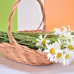 May include: A wicker basket filled with a bouquet of white daisies and green stems. The basket has a woven handle and is sitting on a white surface.