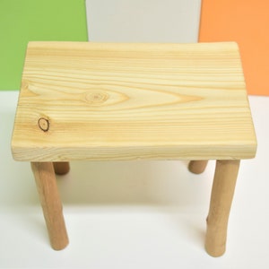 May include: A small, natural wood stool with a rectangular top and four legs. The stool is made of unfinished wood and has a rustic look.