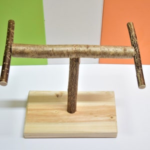 May include: A wooden bird perch with two horizontal branches and a vertical stand. The perch is made of natural wood with a brown bark finish. The stand is made of light wood and has a rectangular shape.