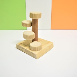 May include: Wooden toy with three platforms and a central post. The platforms are round and the post is cylindrical. The toy is made of natural wood and has a light brown color.