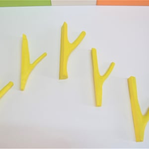 Puede incluir: Cinco ganchos de plástico amarillo con forma de ramas, montados en una pared blanca.