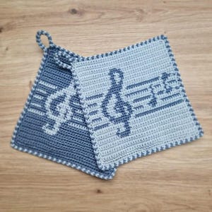 Puede incluir: Dos agarraderas de ganchillo en tonos azules. Una es azul oscuro con un diseño de clave de sol, y la otra es azul claro con una clave de sol y notas musicales. Ambas tienen un lazo para colgar.