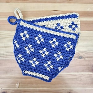Pode incluir: Dois pegadores de panela de crochê em azul e creme com um padrão geométrico. Os pegadores de panela estão dobrados juntos e têm uma alça de laço.