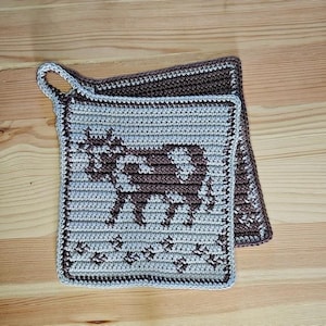 Könnte beinhalten: Zwei gehäkelte Topflappen. Der obere ist hellgrau mit einem braunen Kuhdesign und einer Schlaufe zum Aufhängen. Der Rand ist braun. Der untere Topflappen ist braun. Die Topflappen sind quadratisch und liegen auf einer Holzoberfläche.