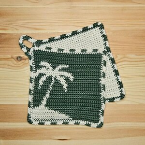 May include: Two handmade crochet potholders. The potholders are square, with a dark green center featuring a white palm tree design. The border and hanging loop are white. The potholders are approximately 8 inches square.