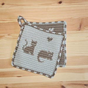 May include: Two crocheted potholders with a cat design. The potholders are light gray with brown cat silhouettes and a brown border. One potholder has a loop for hanging. The potholders are square and appear to be handmade.