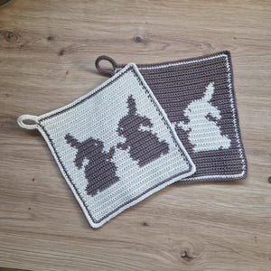 May include: Two crocheted potholders with bunny designs. One is light gray with dark gray bunnies, the other is dark gray with light gray bunnies. Each has a loop for hanging and a white border.