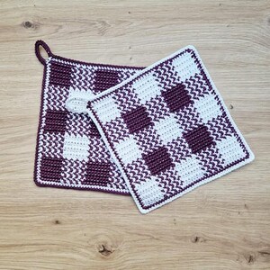 May include: Two handmade crochet potholders with a checkered pattern in white and dark purple. One potholder has a loop for hanging. The potholders are square and rest on a light wood surface.