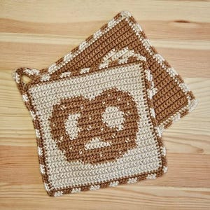 Könnte beinhalten: Zwei quadratische, gehäkelte Topflappen. Die Topflappen sind beige mit einem braunen Brezel-Design in der Mitte. Die Ränder sind mit einem weiß-braunen Muster verziert. Ein Topflappen hat eine Schlaufe zum Aufhängen.