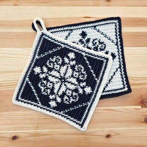 Könnte beinhalten: Zwei gehäkelte Topflappen in einem geometrischen Muster in Schwarz und Weiß. Die Topflappen sind quadratisch und haben einen Schlaufe an einem der Topflappen.
