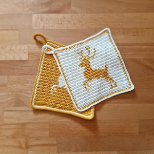 Puede incluir: Dos agarraderas de olla de ganchillo, una amarilla y otra blanca. La agarradera blanca tiene un diseño de reno de ganchillo amarillo.