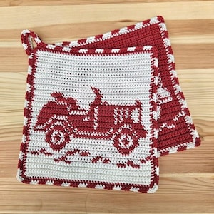 Puede incluir: Juego de agarraderas de ganchillo con un diseño de tractor rojo sobre fondo blanco. Las agarraderas tienen un borde rojo con un borde decorativo blanco y un lazo para colgar. El juego está hecho de hilo suave y texturizado.