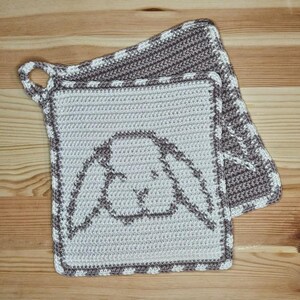 Puede incluir: Dos agarraderas de ganchillo hechas a mano. La agarradera principal es de color gris claro con un contorno de cara de conejo marrón. El borde es marrón con detalles blancos. La segunda agarradera es marrón con detalles blancos. Una tiene un lazo para colgar.