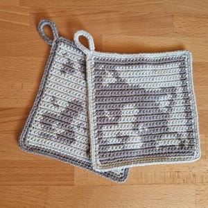 Può includere: Due presine all'uncinetto con un motivo geometrico beige e marrone. Le presine hanno un'ansa a forma di anello.