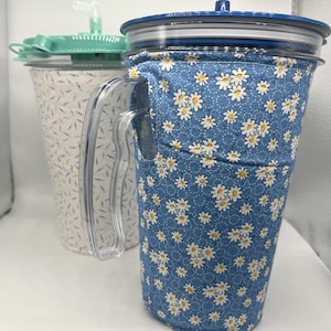 May include: Two insulated tumblers with clear plastic handles and lids. The front tumbler features a blue fabric sleeve with a white daisy pattern. The back tumbler has a white fabric sleeve with a colorful sprinkle pattern and a mint green lid.