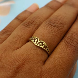 May include: A gold-colored ring with a decorative openwork design. The ring features a curved shape with symmetrical swirling patterns. The ring is worn on a finger, set against a light blue background.