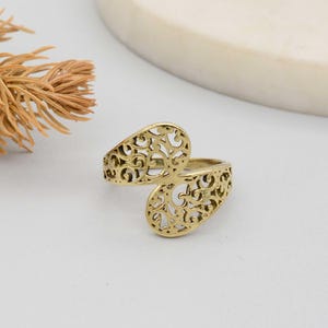May include: A gold-colored ring with an openwork design. The ring features two oval-shaped elements connected by a band, each with intricate cut-out patterns. The ring is set against a neutral background, highlighting its ornate details.
