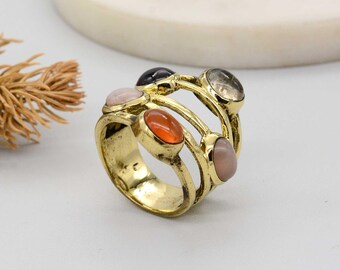 May include: A gold-toned ring featuring multiple oval-shaped gemstones in various colors, including orange, pink, and gray. The ring has a multi-band design, with the gemstones set across the top. The ring is displayed on a white surface.