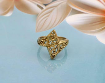 May include: A gold-colored ring with an openwork design. The ring features a central geometric shape with intricate cut-out patterns and a band with similar detailing. The ring is set against a light blue background with floral accents.