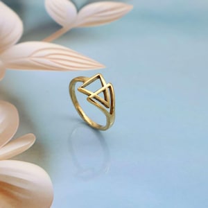 Puede incluir: Un anillo dorado con un diseño geométrico de dos triángulos superpuestos. El anillo está sobre un fondo azul claro, con suaves acentos florales blancos en la esquina superior izquierda. El diseño del anillo es abierto y aireado.