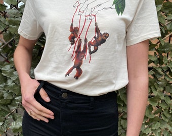 Könnte beinhalten: Ein weißes T-Shirt mit einem Aufdruck einer Hand, die an Fäden hängt, an denen drei Orang-Utans befestigt sind. Die Orang-Utans sind rot und braun und hängen an den Fäden. Die Hand ist schwarz umrandet und die Fäden sind rot.