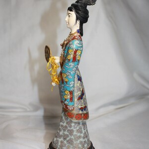 May include: A porcelain figurine of a woman in a blue and white patterned robe with a gold fan. The figurine is on a black wooden base.
