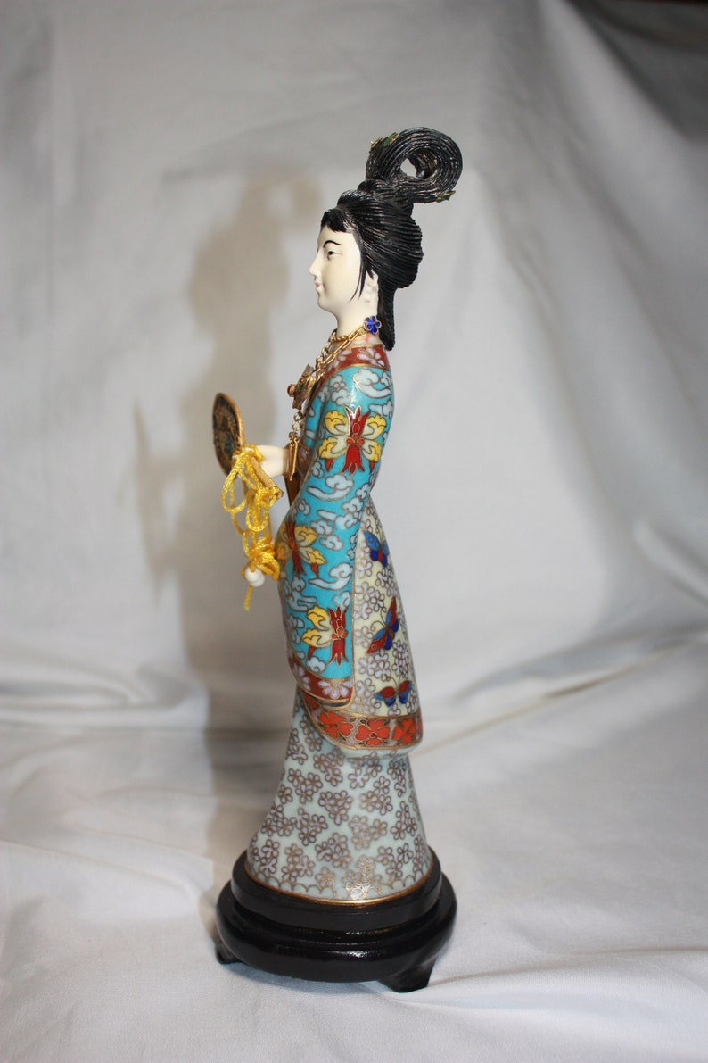 May include: A porcelain figurine of a woman in a blue and white patterned robe with a gold fan. The figurine is on a black wooden base.