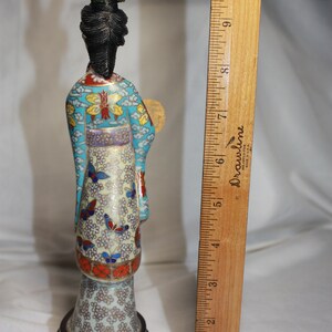May include: A cloisonn&eacute; figurine of a woman in a blue, white, and gold robe with a black hairpiece and a fan. The figurine is standing on a black base.