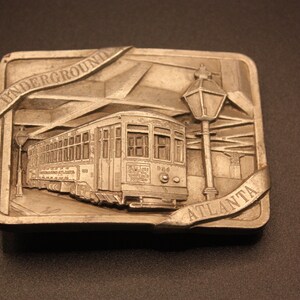 May include: A silver belt buckle with a raised design of a streetcar, a street lamp, and the words "UNDERGROUND ATLANTA".