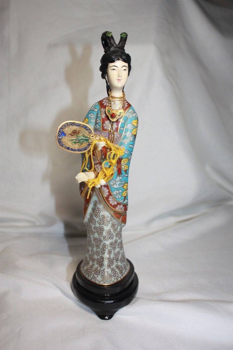 May include: A porcelain figurine of a woman in a blue, red, and gold patterned robe holding a fan. The figurine is standing on a black wooden base.