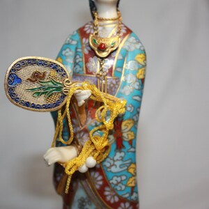 May include: A porcelain figurine of a woman in a blue, red, and gold patterned robe. She is holding a fan with a floral design and a long string of beads.