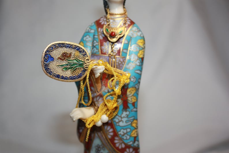 May include: A porcelain figurine of a woman in a blue, red, and gold patterned robe. She is holding a fan with a floral design and a long string of beads.