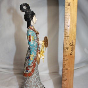 May include: A porcelain figurine of a woman in a blue and gold patterned robe, holding a golden fan and a string of beads. The figurine is standing on a black base.